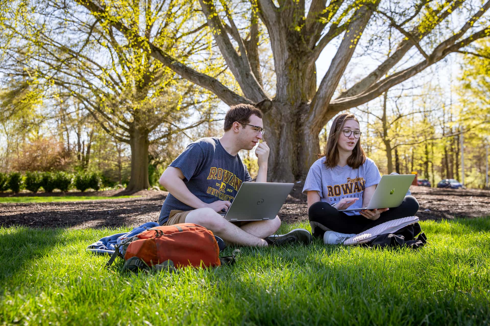Two Rowan students work on their laptops while sitting in the grass.