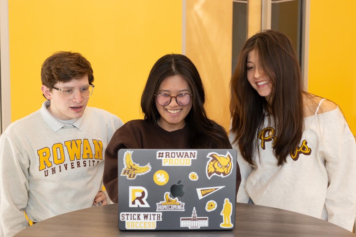 Three Rowan students smile while looking at a laptop covered in Rowan University stickers.