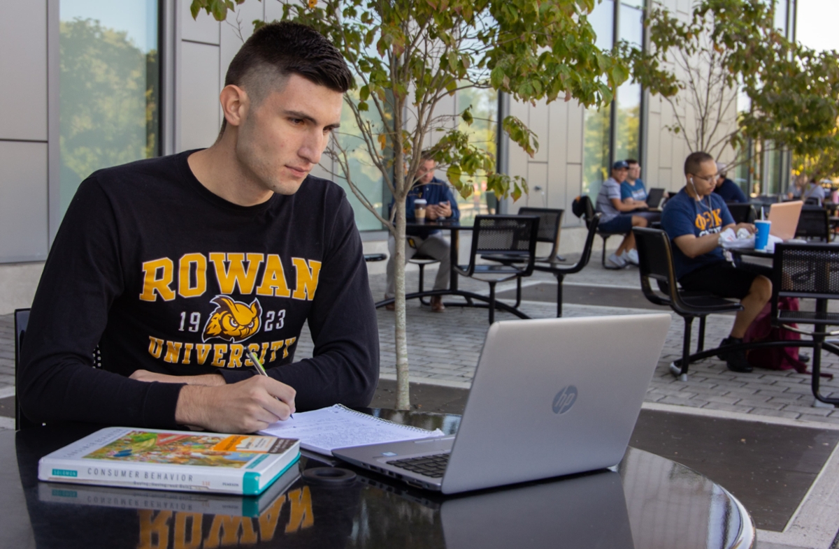 A Rowan student sits at a table while working on their laptop on Rowan's main campus in Glassboro, NJ. 
