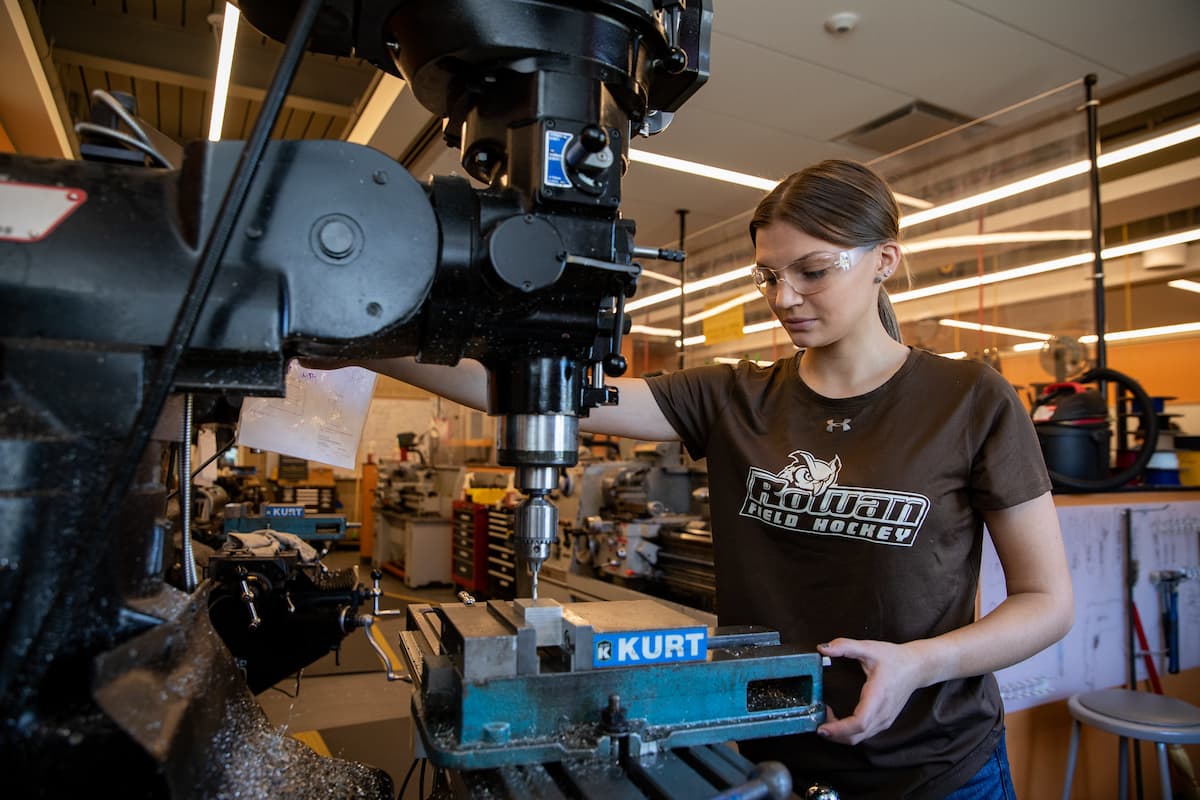 student wearing safety-goggles and rowan t-shirt operating a large industrial drill in a workshop