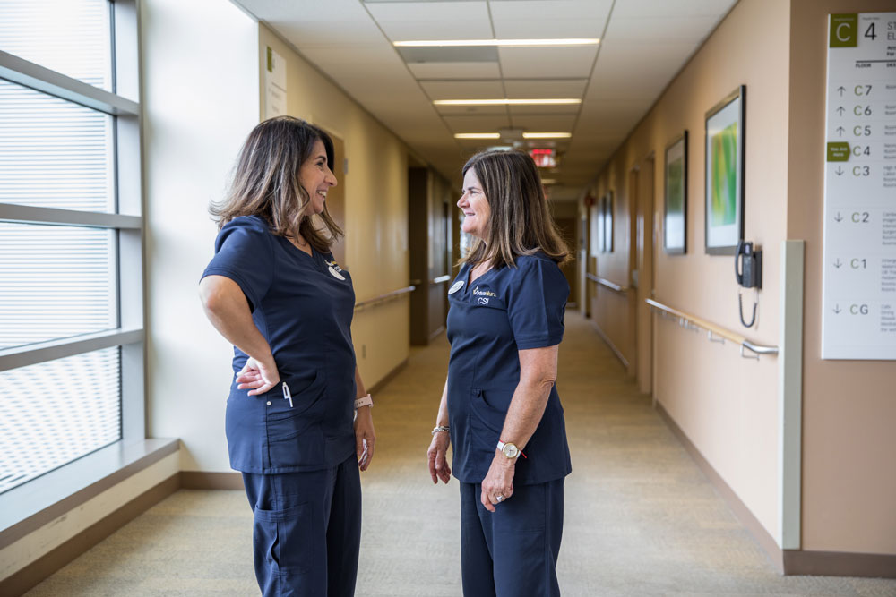 Two nurses smile while talking to each other in a hallway.
