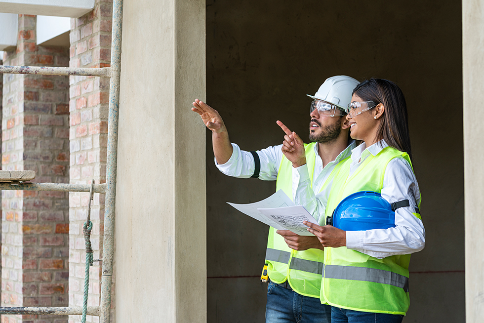 Two professionals stand together in construction gear while pointing at something.