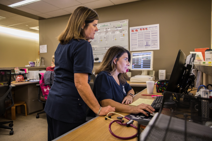 Two nurses work together on a computer at a desk in a healthcare setting.