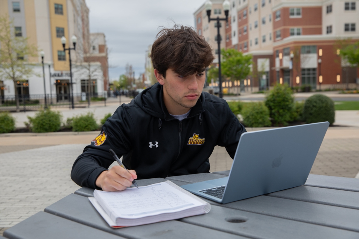 A Rowan student sits at a picnic table while working on their laptop.