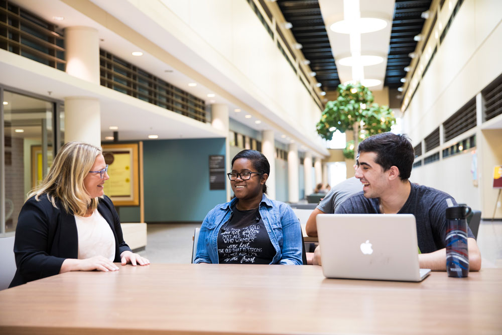 A professor sits with two students at a table while they are smiling and laughing.