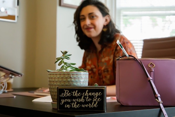 A teacher sits at her desk behind a sign that reads, Be the change you wish to see in the world.