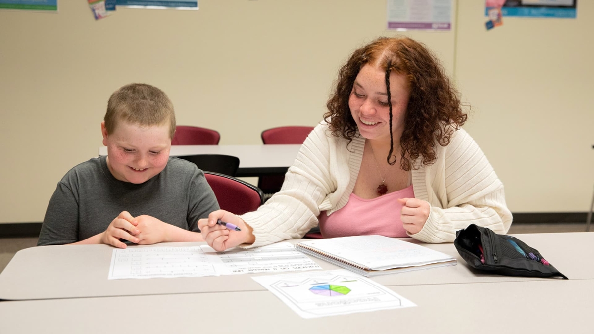 A Rowan student sits at a desk with a child while smiling and helping them.