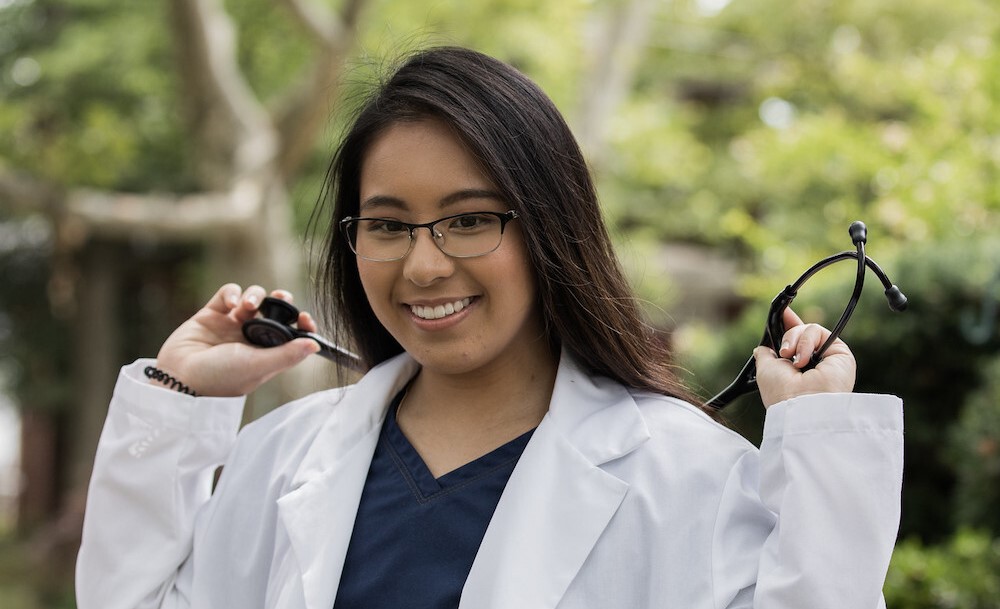 A Rowan student smiles in a white coat with a stethoscope around their neck.