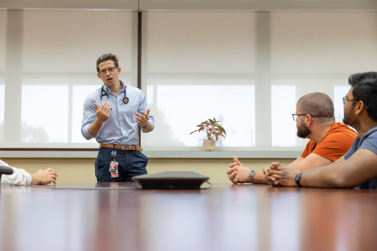 A medical student stands and speaks with a group sitting around a table.