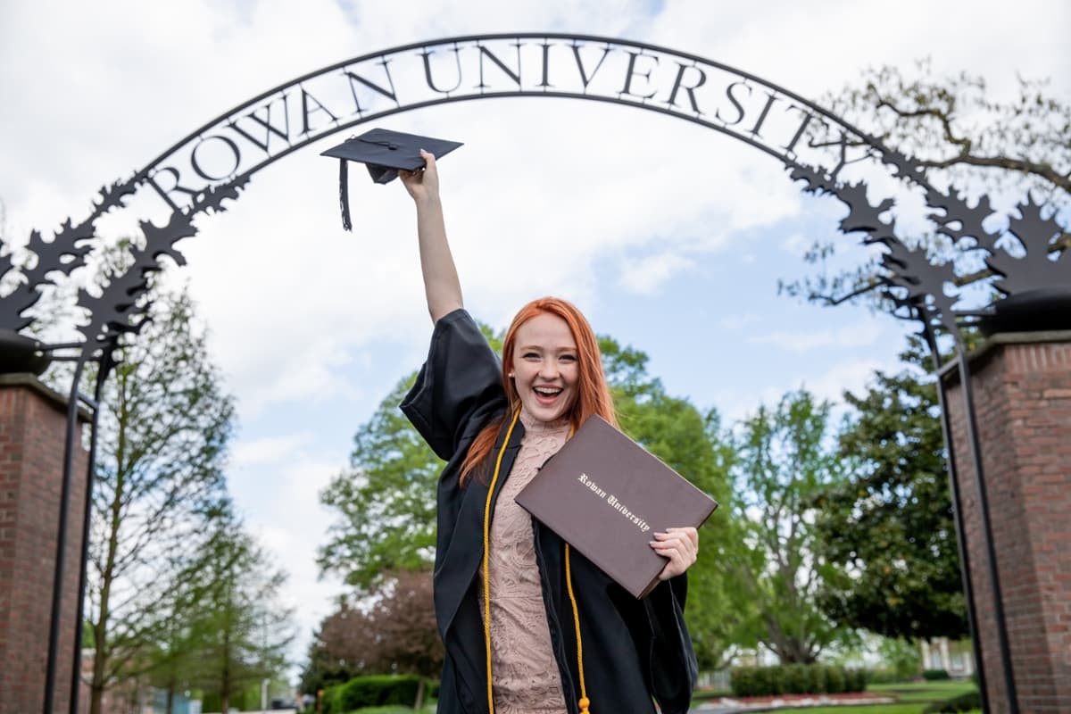 A Rowan student smiles while wearing a graduation gown and holding a graduation cap and a degree cover under the Rowan University arch.